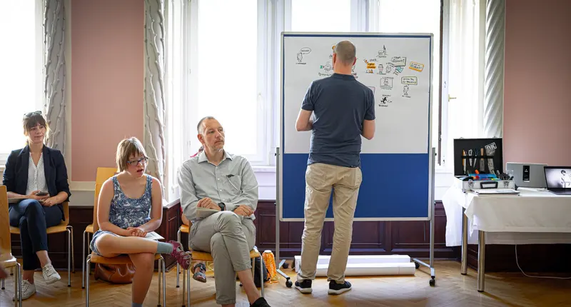 Person zeichnet an Flipchart, zwei Personen sitzen daneben.