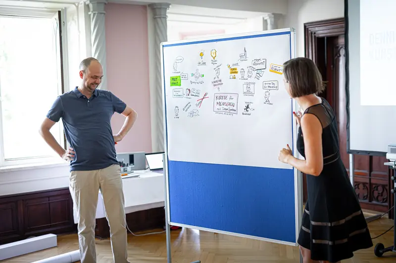 Person zeichnet an Flipchart, weitere Person steht daneben.