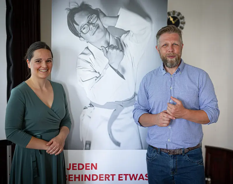 Zwei Personen stehen lächelnd vor einem Plakat.