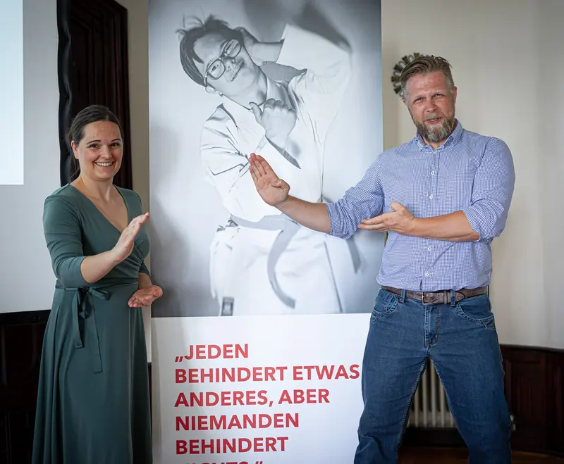 Zwei Personen sprechen gestikulierend vor einem Plakat.