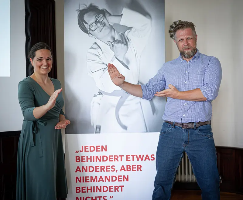 Zwei Personen unterhalten sich gestikulierend vor einem Plakat.
