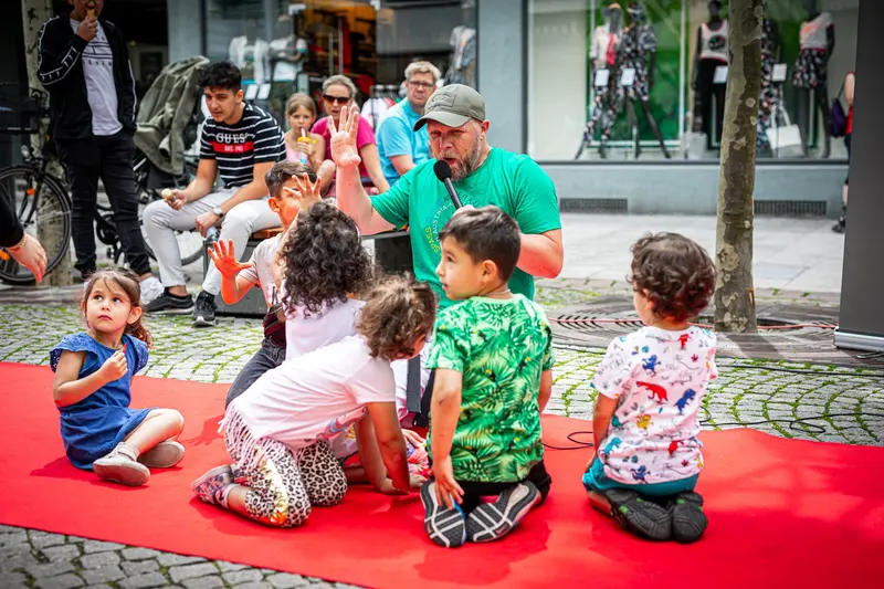 Trainer mit Mikrofon spricht mit sitzenden Kindern auf rotem Teppich