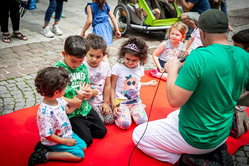 Trainer sitzt mit Kindergruppe im Kreis auf rotem Teppich