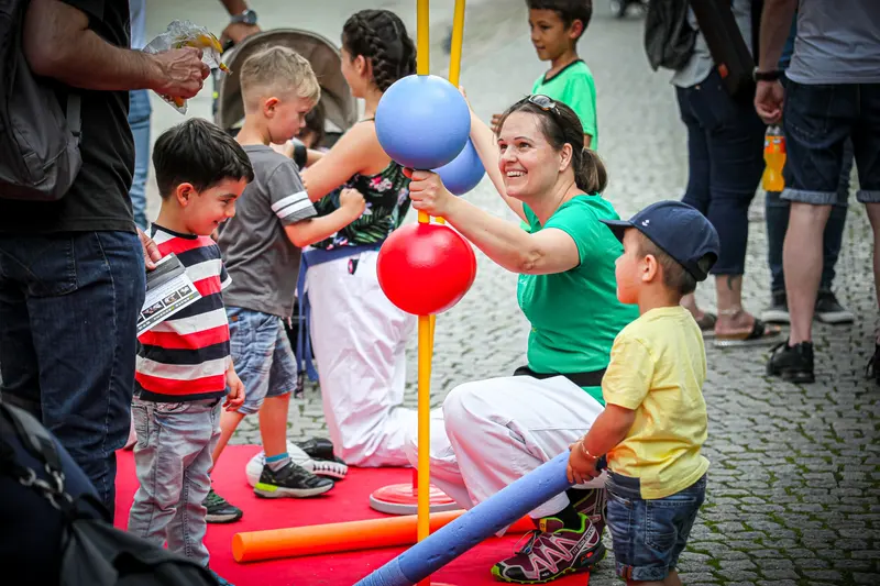 Person hält Kindern einen Luftballon hin, im Hintergrund Fußgängerzone