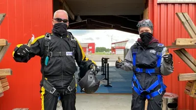 Zwei Personen in Fallschirmspringer-Ausrüstung stehen vor einem Hangar