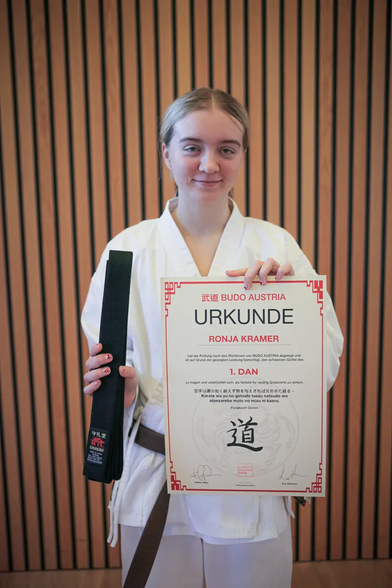 Person im Karategi hält lächelnd Urkunde und schwarzen Gürtel, Porträt