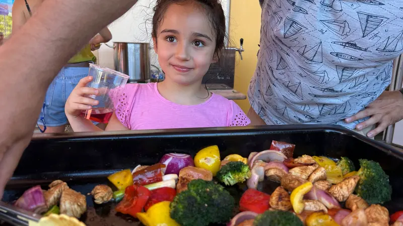 Kind hält lächelnd ein Getränk vor dem Grill mit Gemüse