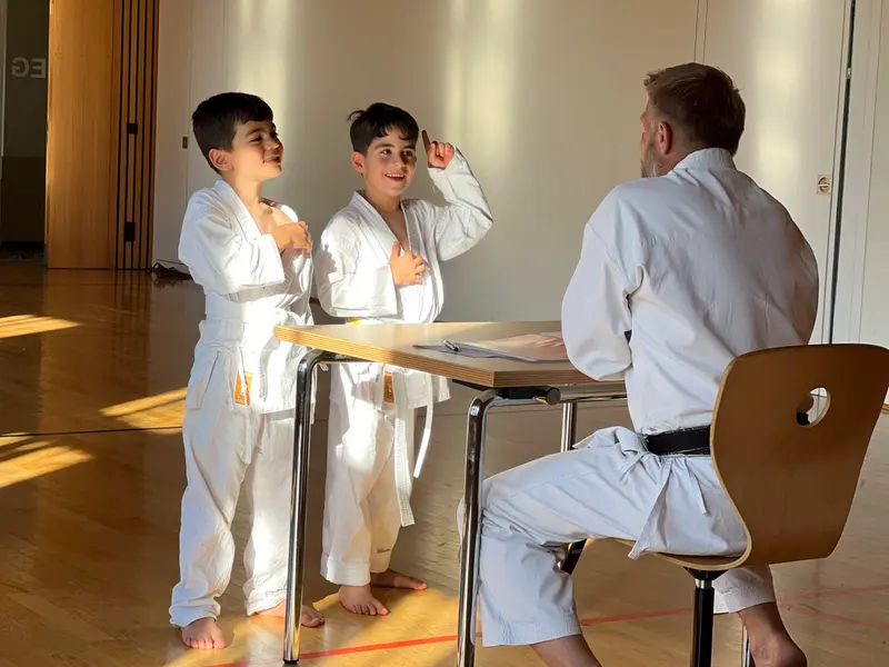 Zwei Kinder sitzen am Tisch, Prüfer daneben