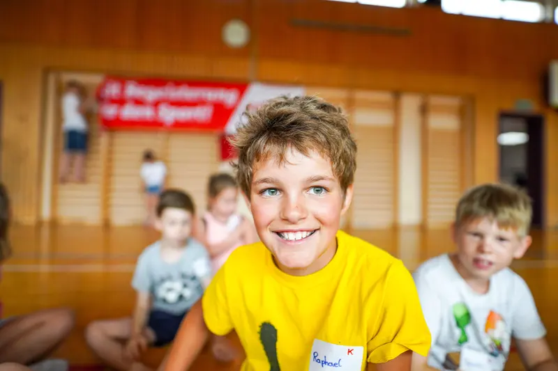 Gruppe Kinder in Turnhalle, Junge im gelben Shirt lächelt in die Kamera