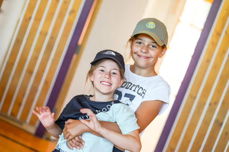 Zwei Kinder mit Kappen posieren lachend, eine winkt in die Kamera