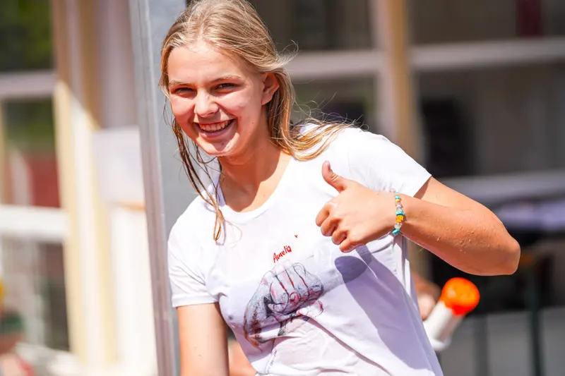 Person im weißen Shirt zeigt lächelnd Daumen hoch, nass von Wasserspiel