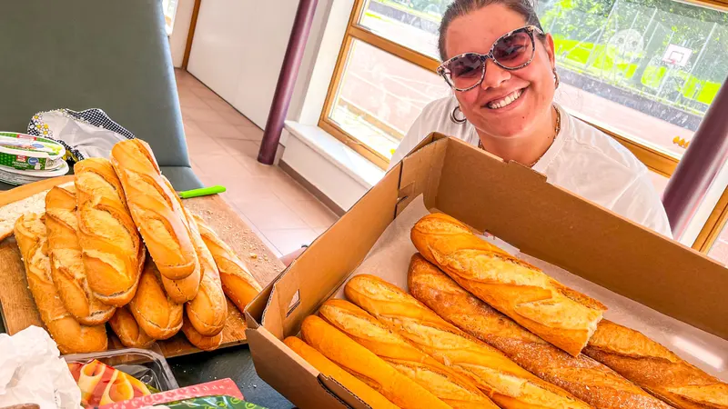 Person mit Sonnenbrille hält Karton mit Baguettes vor Fenster