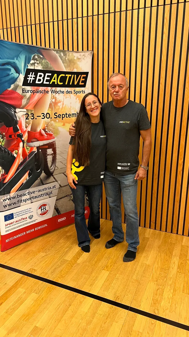 Person und Trainer im schwarzen Shirt posieren lächelnd vor Fitness-Plakat