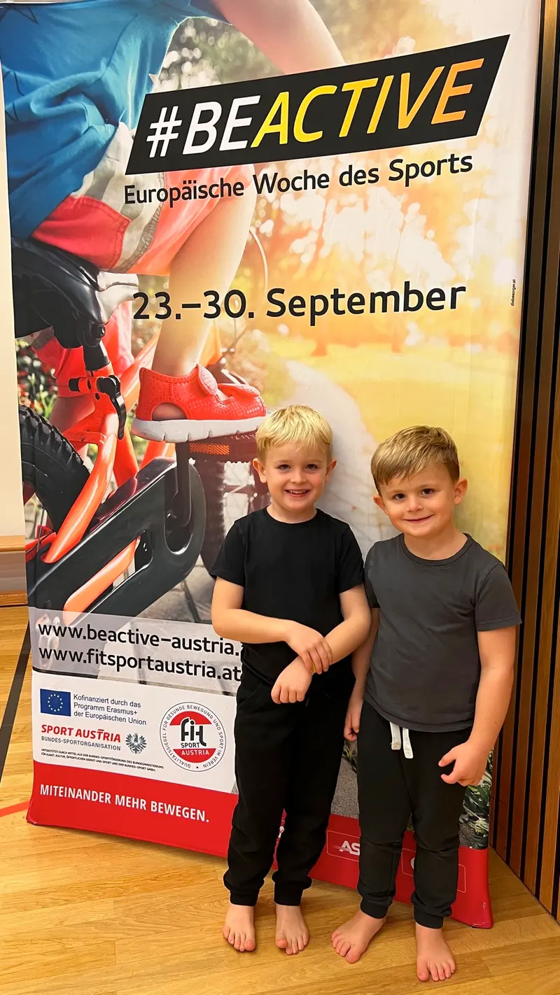 Zwei Kinder posieren lächelnd vor #BeActive-Plakat mit roten Sportschuhen