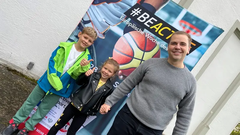 Kinder und Erwachsener posieren lächelnd vor Basketball-Plakat