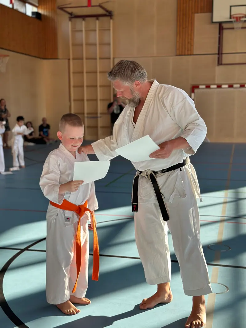 Prüfer überreicht einem Kind mit orangem Gürtel ein Blatt