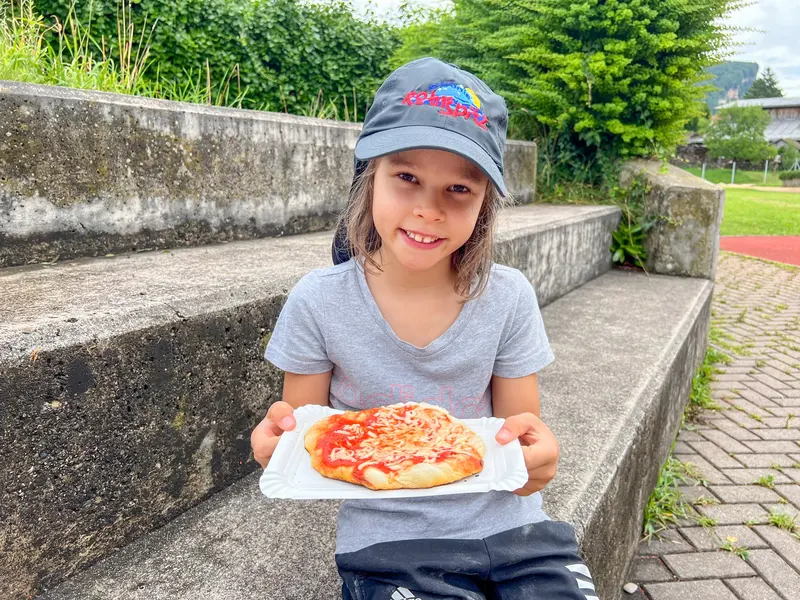Kind mit Kappe sitzt auf Treppe und hält Pizzastück