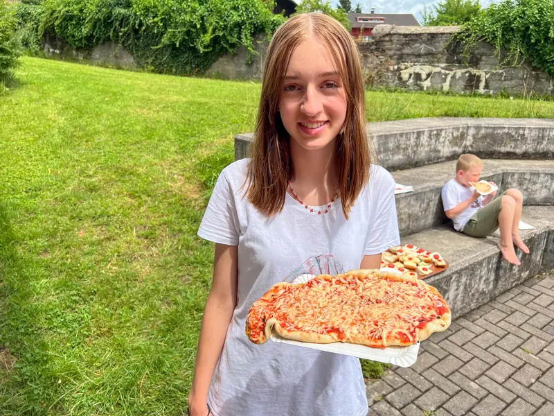 Kind hält lange, selbstgemachte Pizza auf einem Blech im Freien