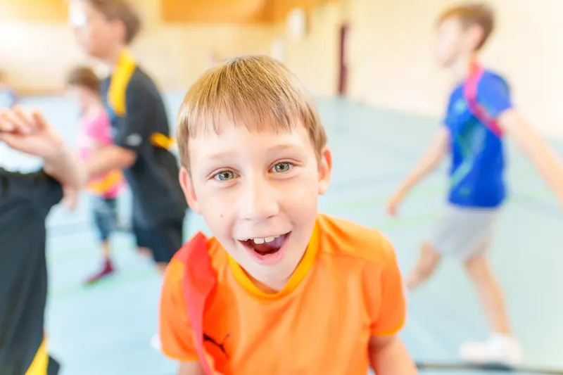 Kind in orangem Shirt lacht, im Hintergrund läuft weiteres Kind
