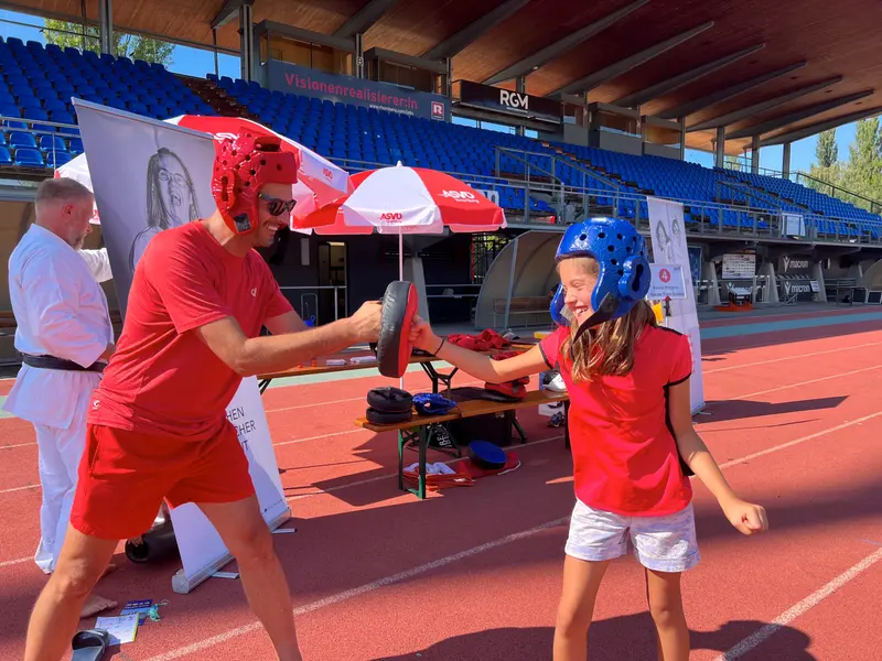 Erwachsener hält Boxpratzen für Kind mit Helm vor Sonnenschirm