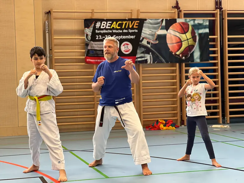 Zwei Personen im Karategi üben Kicktechnik vor Sportbanner