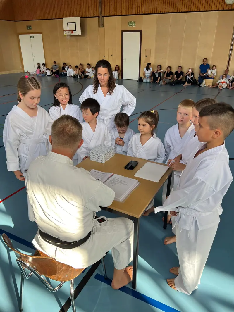 Trainer sitzt am Tisch, Kinder im Karategi stehen darum