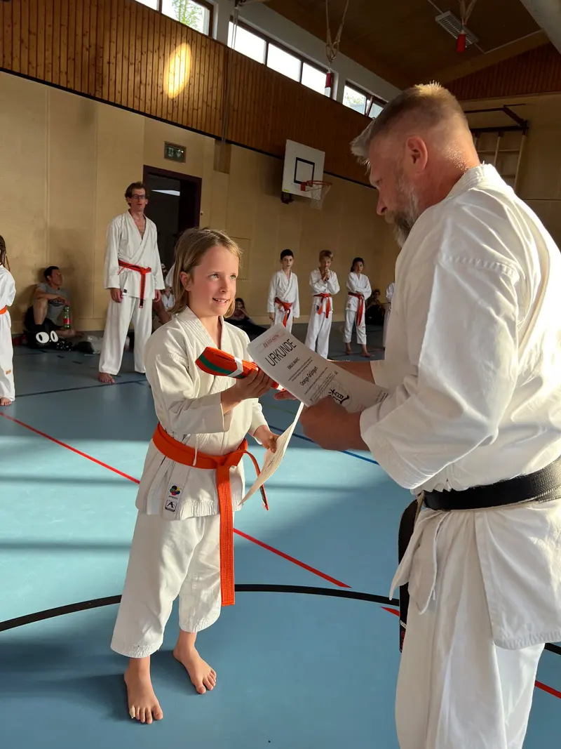Trainer überreicht Kind mit orangem Gürtel eine Urkunde