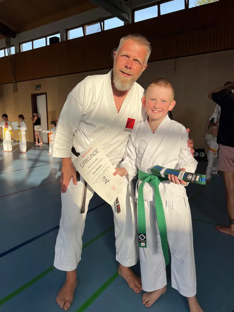 Trainer und Kind lächeln gemeinsam, Kind hält grünen Gürtel