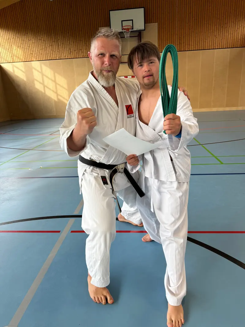 Trainer und Person im Karategi stehen nebeneinander, Person hält grünen Gürtel