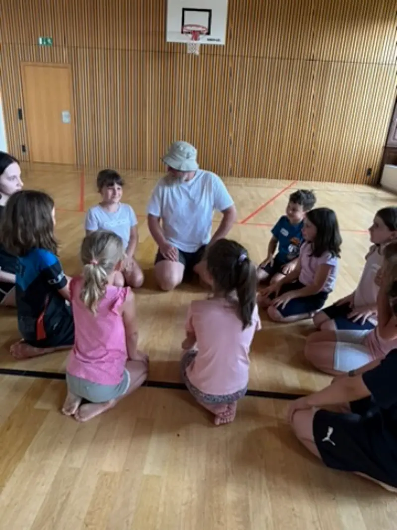 Gruppe Kinder sitzt im Kreis auf einer Matte in der Sporthalle