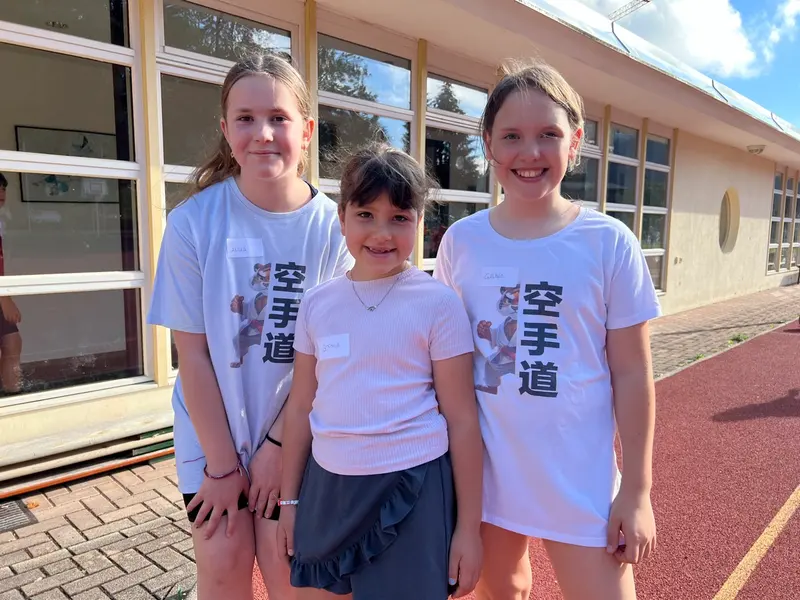Drei Kinder in weißen T-Shirts stehen lächelnd vor einem Gebäude