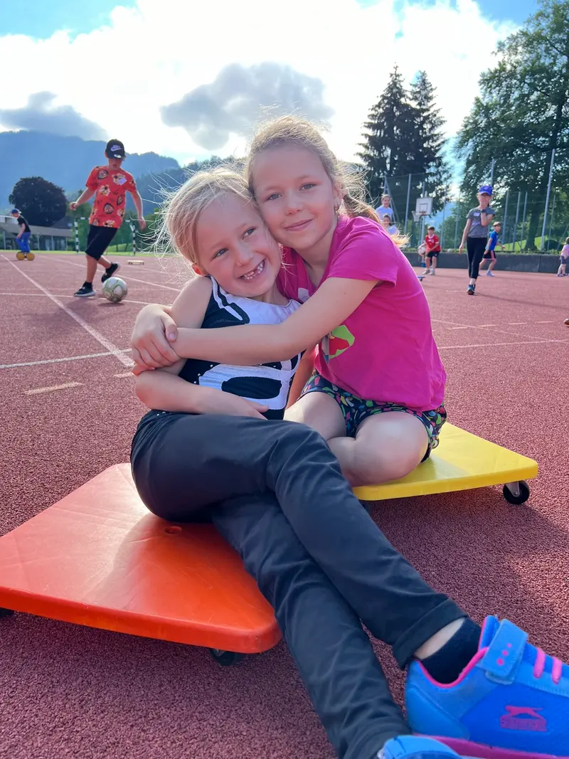 Zwei Kinder sitzen umarmt auf einem Rollbrett auf einer Laufbahn