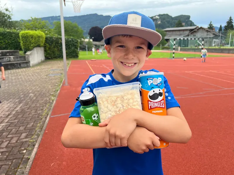 Kind mit Kappe hält Trinkflasche und Snackpackungen im Arm