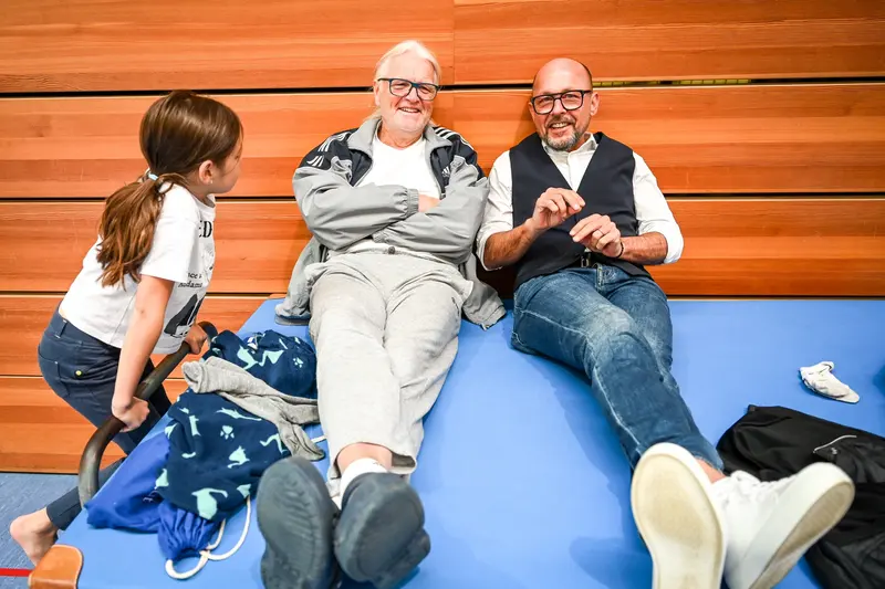 Kind sitzt zwischen zwei Erwachsenen auf blauer Turnmatte