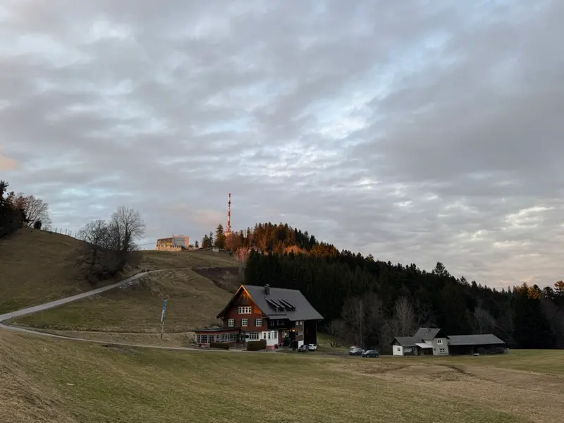 Berghütte auf Wiese mit Sendemast im Hintergrund, bewölkter Himmel