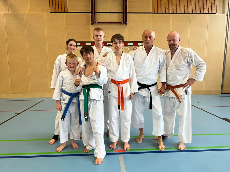 Sechs Personen in Karategi mit verschiedenfarbigen Gürteln stehen in Turnhalle