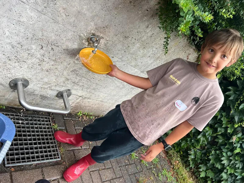 Kind hält gelbe Schüssel unter einen Wasserhahn im Freien