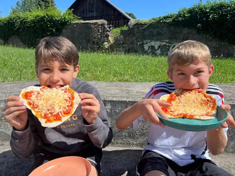Zwei Kinder essen draußen jeweils ein Stück Pizza