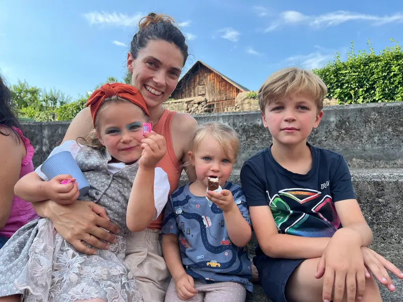 Erwachsene Person umarmt lachend zwei Kinder im Freien.