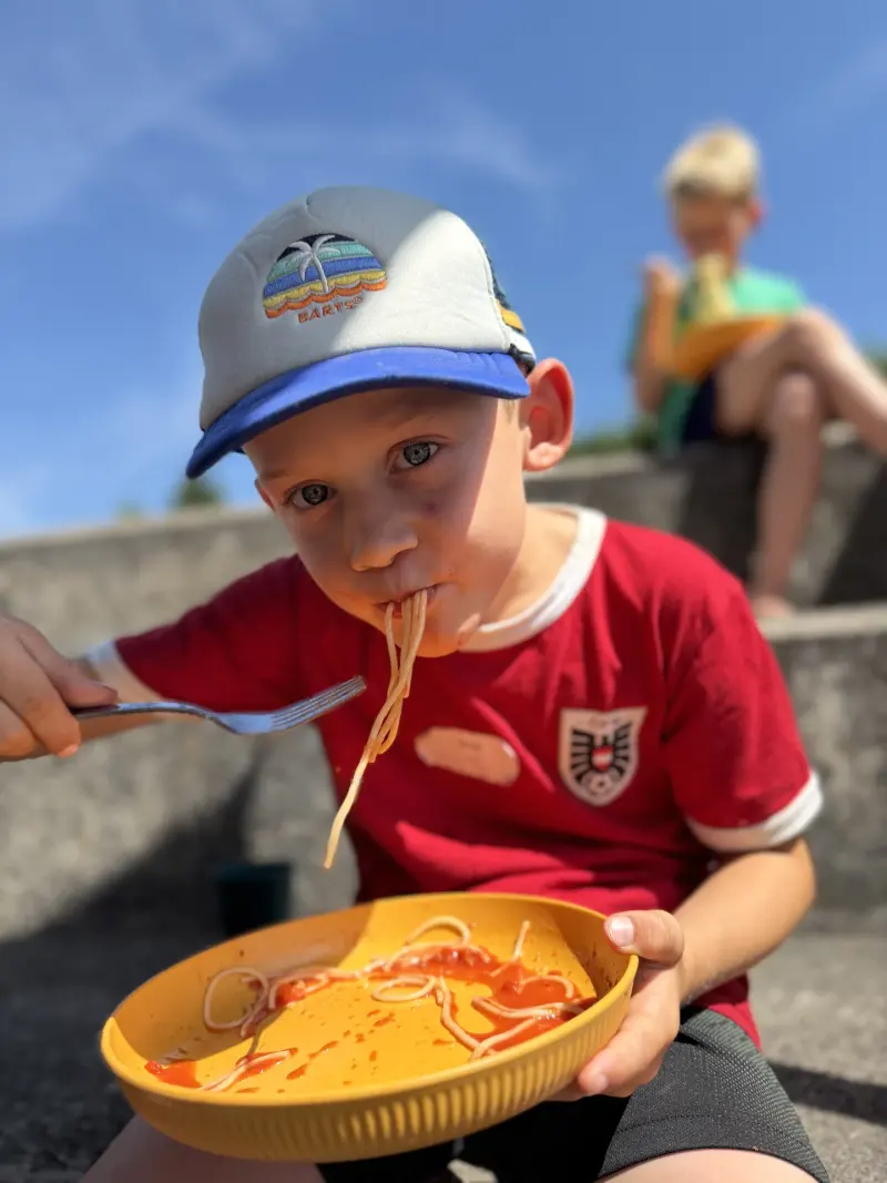 Kind mit Kappe isst Spaghetti im Freien