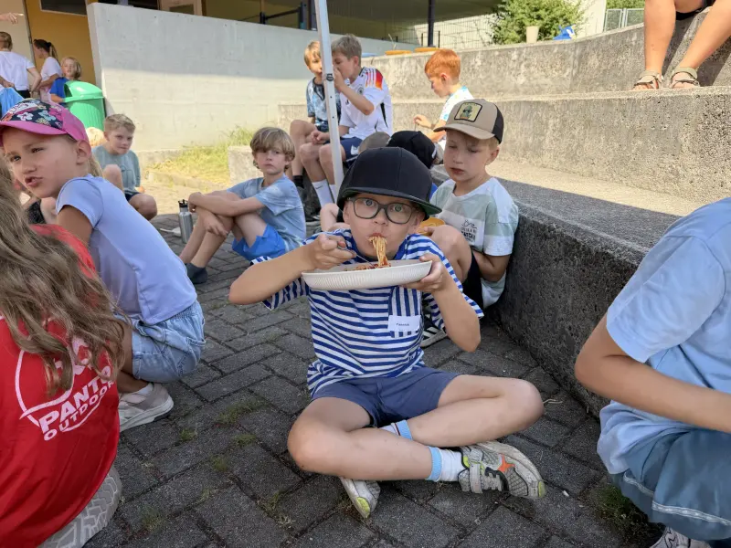 Kind sitzt inmitten einer Gruppe und isst Nudeln